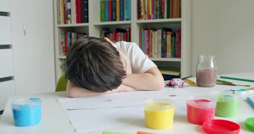 Child Resting Head on Table with Painting Supplies