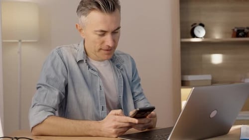 Kaukasischer Mann benutzt Handy zum Telefonieren, während er mit Laptop am Schreibtisch sitzt