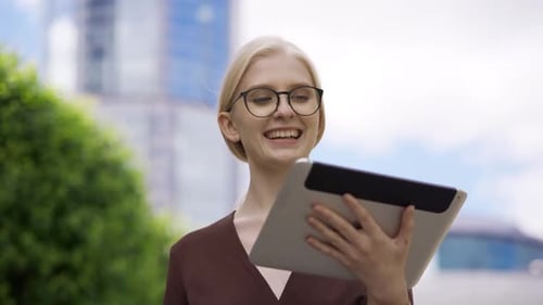 A Young Blonde Woman in Glasses Scrolls the Tablet with Interest in the Street