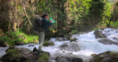 Female Traveler with a Backpack Drinking Water in Nature in the Forest Near a Mountain River
