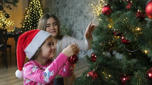 Mom and Daughter in a Santa Hat Decorate the Christmas Tree with Balls Preparing