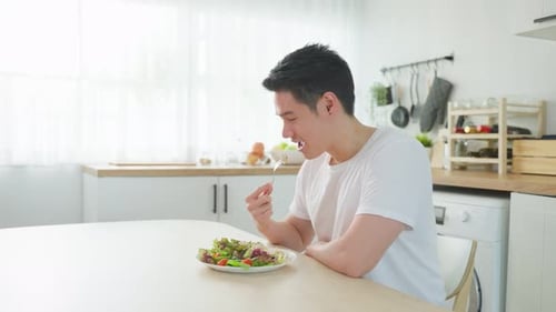 Asian attractive handsome male eating green salad in kitchen at home.