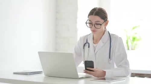 Female Doctor Browsing Internet on Smartphone while using Laptop in Office
