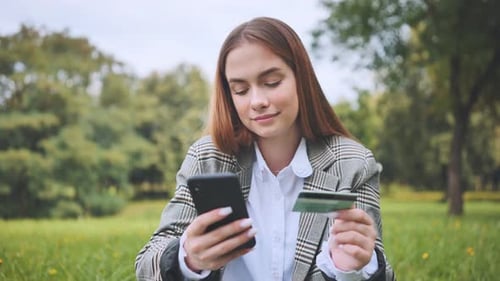 A Young Girl Makes a Purchase with Her Phone and Bank Card in the Park Sitting on the Grass