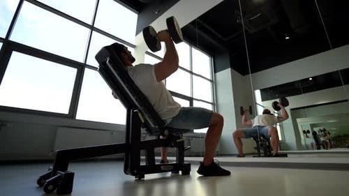 Mature Man Lifting Up Big Dumbbells To Pump Up His Biceps, Sitting on Sport Gym Bench Indoor of