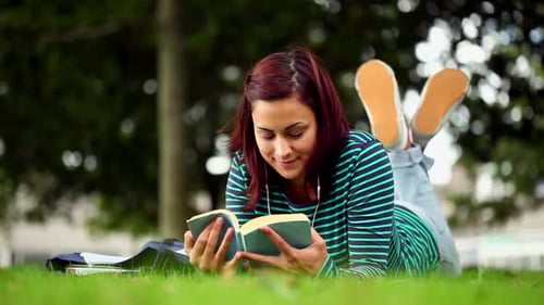 Hübsche Studentin liegt auf Gras und liest ein Buch und hört Musik