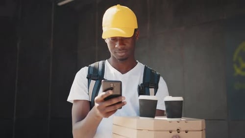 Afrikanischer Zusteller checkt sein Telefon beim Tragen einer Schachtel mit Pizza und