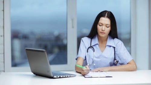 Female Doctor Writing at Desk with Laptop
