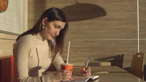 Young Woman in a Cafe Makes Notes in a Notebook and Drinks Juice