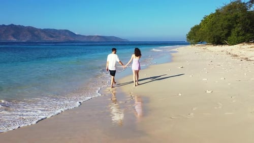 Young romantic couple holding hands while walking along the tropical white sand beach. Mountains in