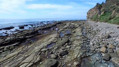 Tracking shot of a young man running on a rocky ocean beach shoreline.