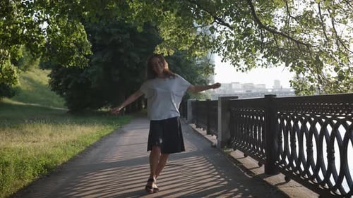 uma mulher com roupas casuais está dançando alegremente em um parque da cidade perto de uma treliça a céu aberto