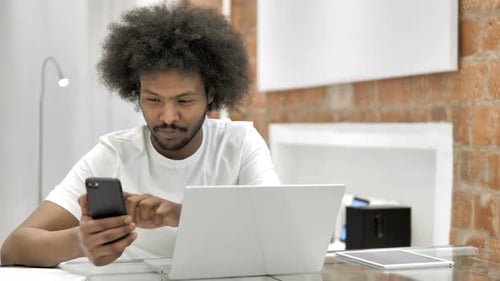 Young Adult Using Smartphone at Desk with Laptop
