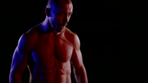 Ein muskulöser Mann mit einem nackten Torso senkt die Arme und schlägt dann vor einem dunklen Hintergrund