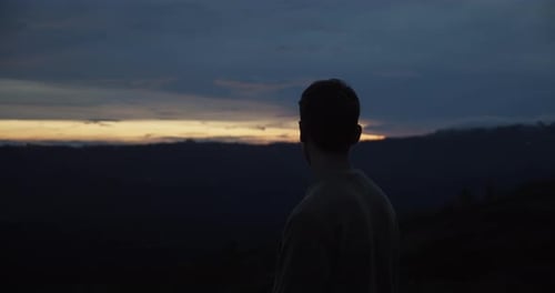 Statische Aufnahme eines weißen Mannes auf dem Gipfel des Hügels während des Sonnenuntergangs, der sich im Dunkeln umschaut
