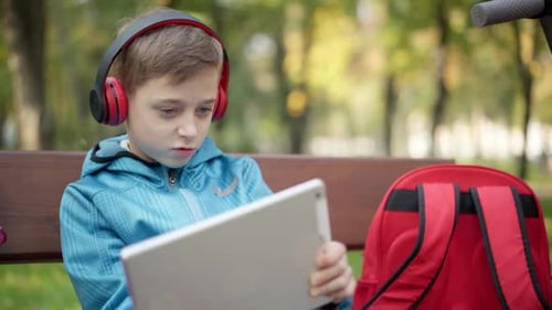 Engrossed Caucasian Boy in Headphones Gaming Online on Tablet Sitting in Autumn Park Outdoors