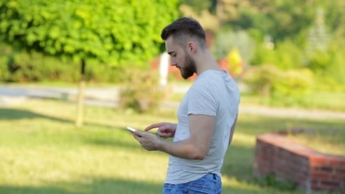Young Man Standing In The Park With Mobile Phone
