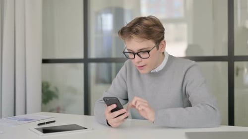 Attractive Young Man Celebrating Success on Smartphone in Office