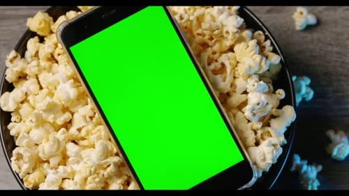 Smart phone with green touch screen and popcorn in a big bowl on the table.