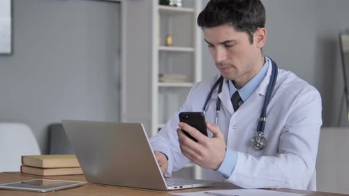 Doctor Using Phone and Laptop at Desk Indoors
