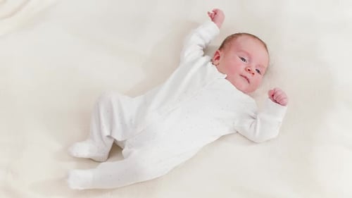 Top View on Adorable Newborn Baby in White Bodysuit Lying on Bed and Looking on Bright Sun