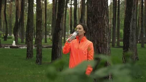 Sportive woman drinks water from fitness bottle after outdoor running training in the park.