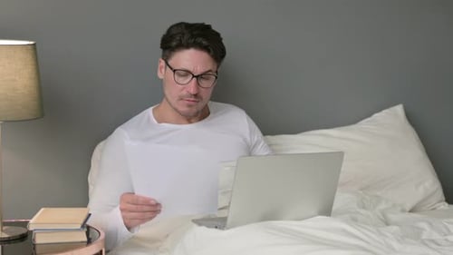 Man Works From Bed With Laptop and Documents