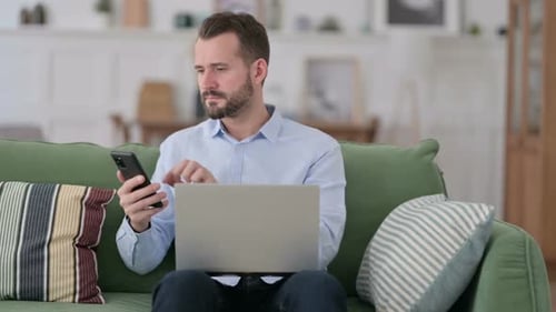 Man Using Smartphone and Laptop on Couch