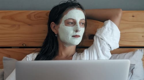 Woman Relaxing in Bed with Beauty Mask and Laptop