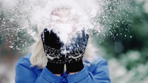 Pretty Woman Blowing Snow From Hands with Mittens To Camera on Winter Forest Background