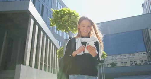 Chica en ropa casual con mochila de pie cerca del edificio de oficinas y usa su móvil