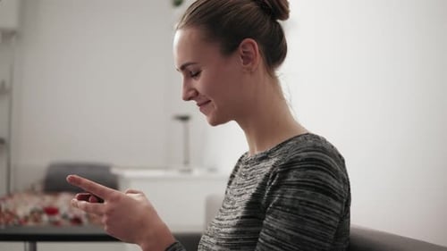 Closeup View of a Smiling Young Woman Sitting on the Couch at Home Looking at Phone and Typing a