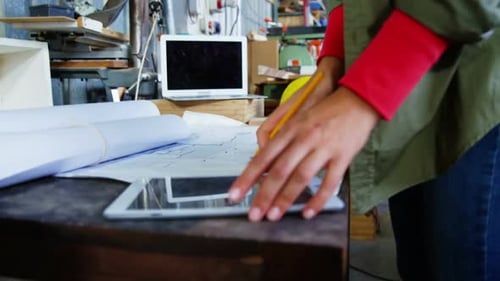 Woman Reviews Blueprints with Tablet in Workshop