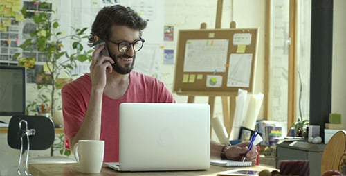 Smiling Man Talks on Phone While Using Laptop