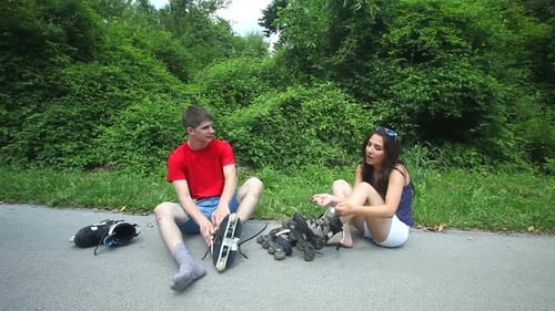 Young Woman And Man Sitting On Track, Putting Their Rollerblades On Their Feet. 1
