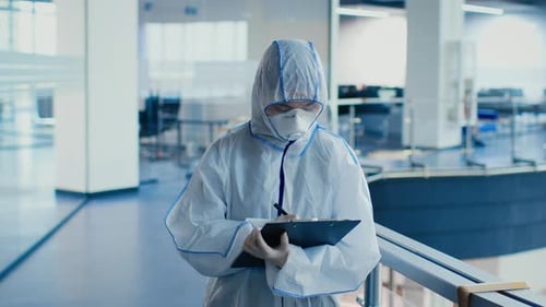 Doctor Wearing Hazmat Suit And Mask Taking Notes In Clinic