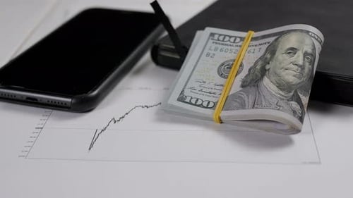 A Stack of Hundreddollar Bills on a Light Wooden Table with Financial Charts