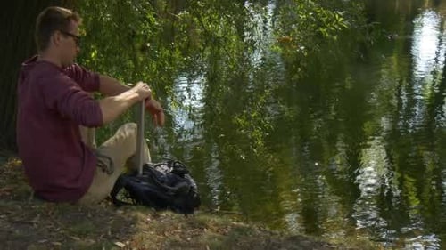 Young Adult Works on Laptop by Riverside Tree