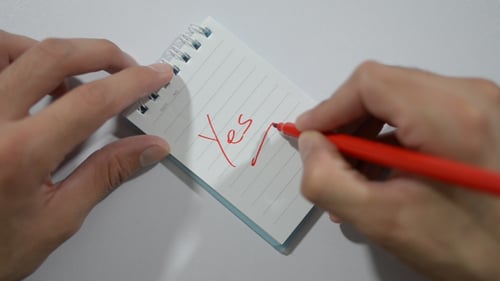 Hands Write 'Yes' in Red Pen on Notepad