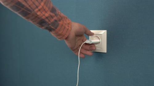 A Man Pulls Out a Phone Charger From a Wall Outlet. Rosette in the Wall with Dark Blue Wallpaper