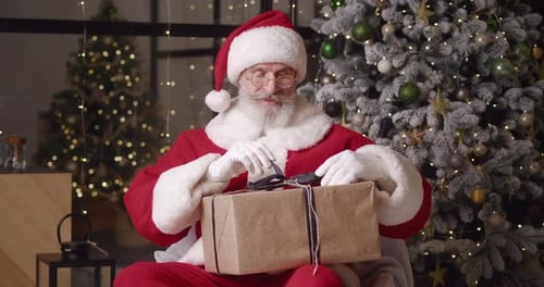 Santa Claus Unwrapping Present by Christmas Tree