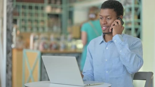 Young African Man with Laptop Talking on Smartphone in Cafe