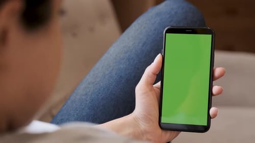 Young woman sitting on a couch and using smartphone with green screen