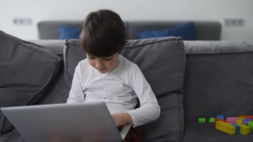 Kleiner süßer Junge benutzt Laptop, der alleine auf dem Sofa sitzt