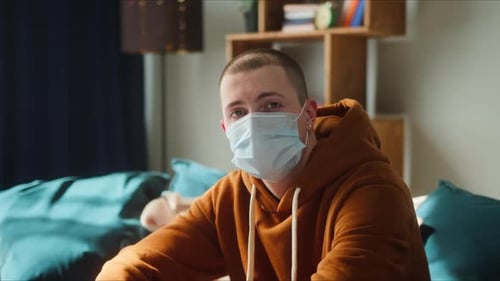 Porträt eines jungen amerikanischen Mannes, der auf dem Sofa sitzt und eine medizinische OP-Maske trägt, um das
