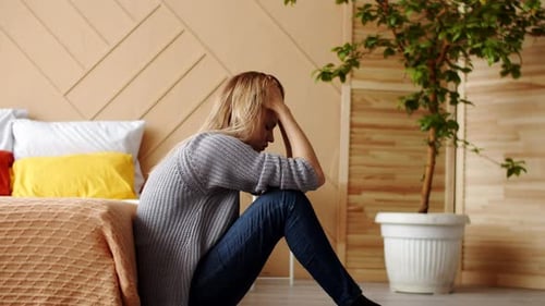 Woman is Sitting on Floor By Bed Holding Head and Swaying From Stress of Life Side View