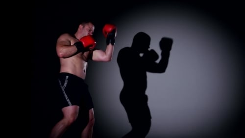 Shirtless Boxer Practices Punches with Shadow in Dark Gym