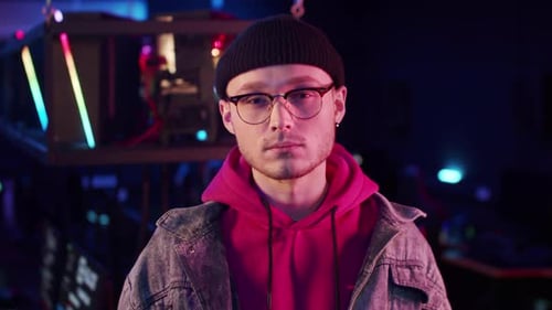 A Young Gamer Guy in a Hoodie Denim Jacket Knitted Hat and Glasses Stares Intently at the Camera