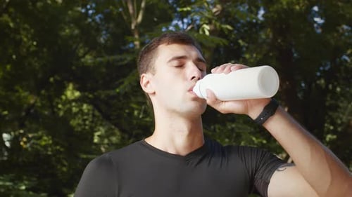 Tracking Shot Portrait of Young Guy Athlete Drinking Water in Green Park Taking Break After Outdoors
