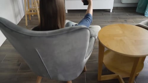 Woman is Sitting in a Chair Watching TV with a Green Screen Switching Channels with a Remote Control